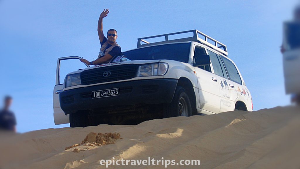 Jeep on the edge of the Sahara dunes with man waving. Part of our Adventurous Three Days Epic Sahara Tour In Amazing Tunisia With Hidden Gems.