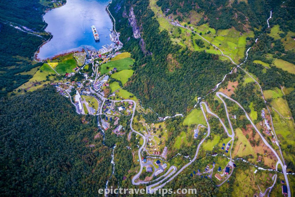 Aerial photo of Geiranger village, fjord, winding roads, cruise ship, and forests around.